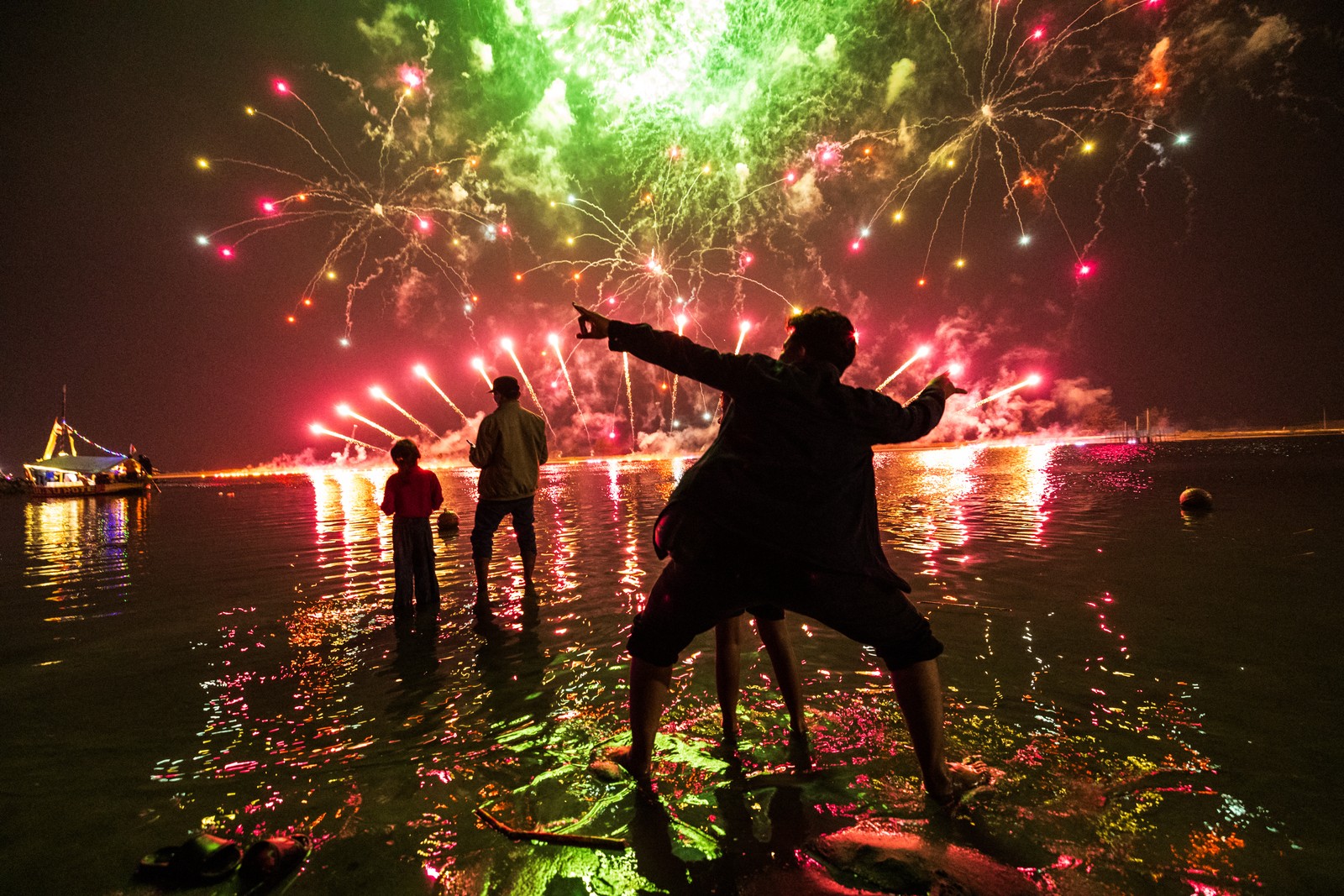 People stand in shallow water, gesturing, as they enjoy a fireworks show.