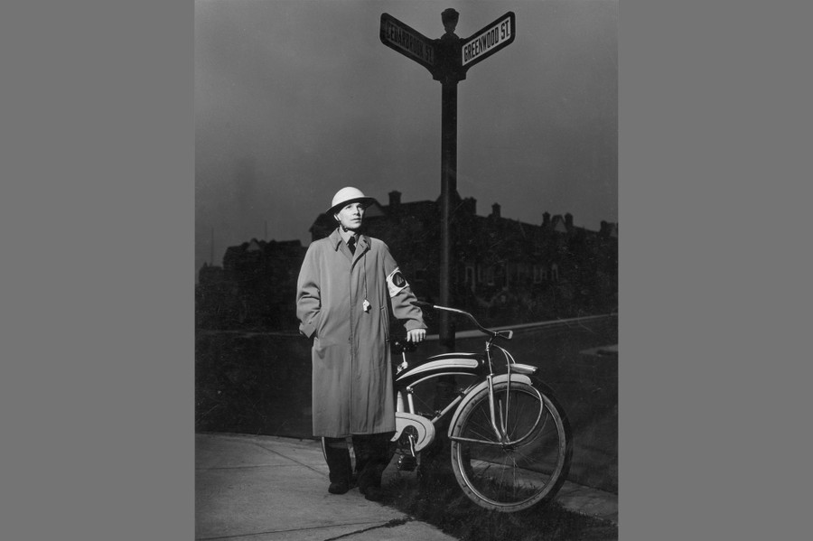 A Civil Defense air-raid warden stands beside a street sign and his bicycle.