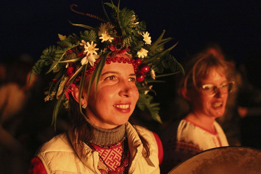 A woman wearing traditional clothing and a floral wreath is illuminated by a nearby bonfire.