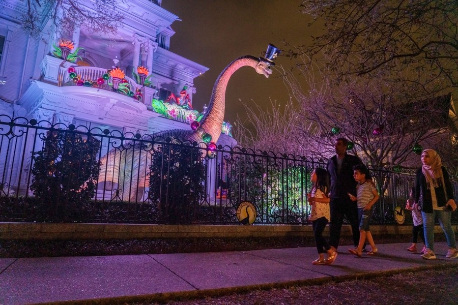 People walk past a large dinosaur standing in front of a decorated house.
