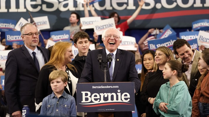 Bernie Sanders, standing at a lectern, surrounded by his family, laughs as he looks onto a crowd.