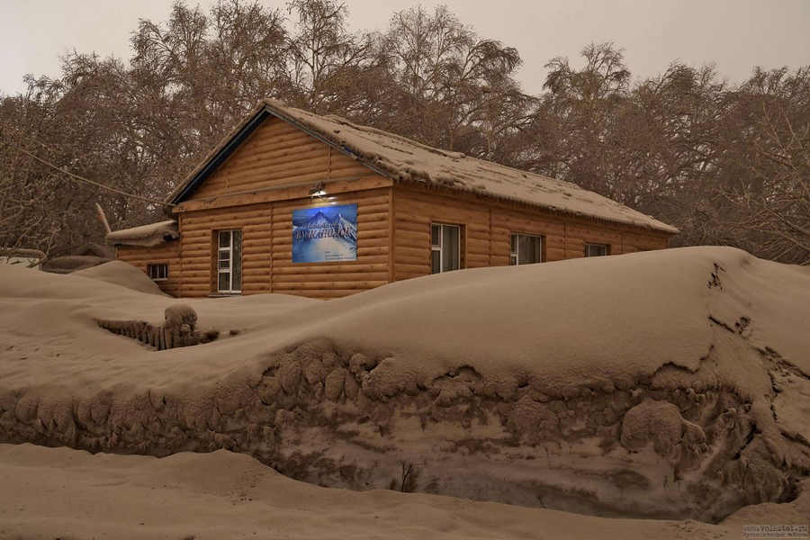 A gray blanket of ash covers snowbanks, the roof of a wooden building, and trees.