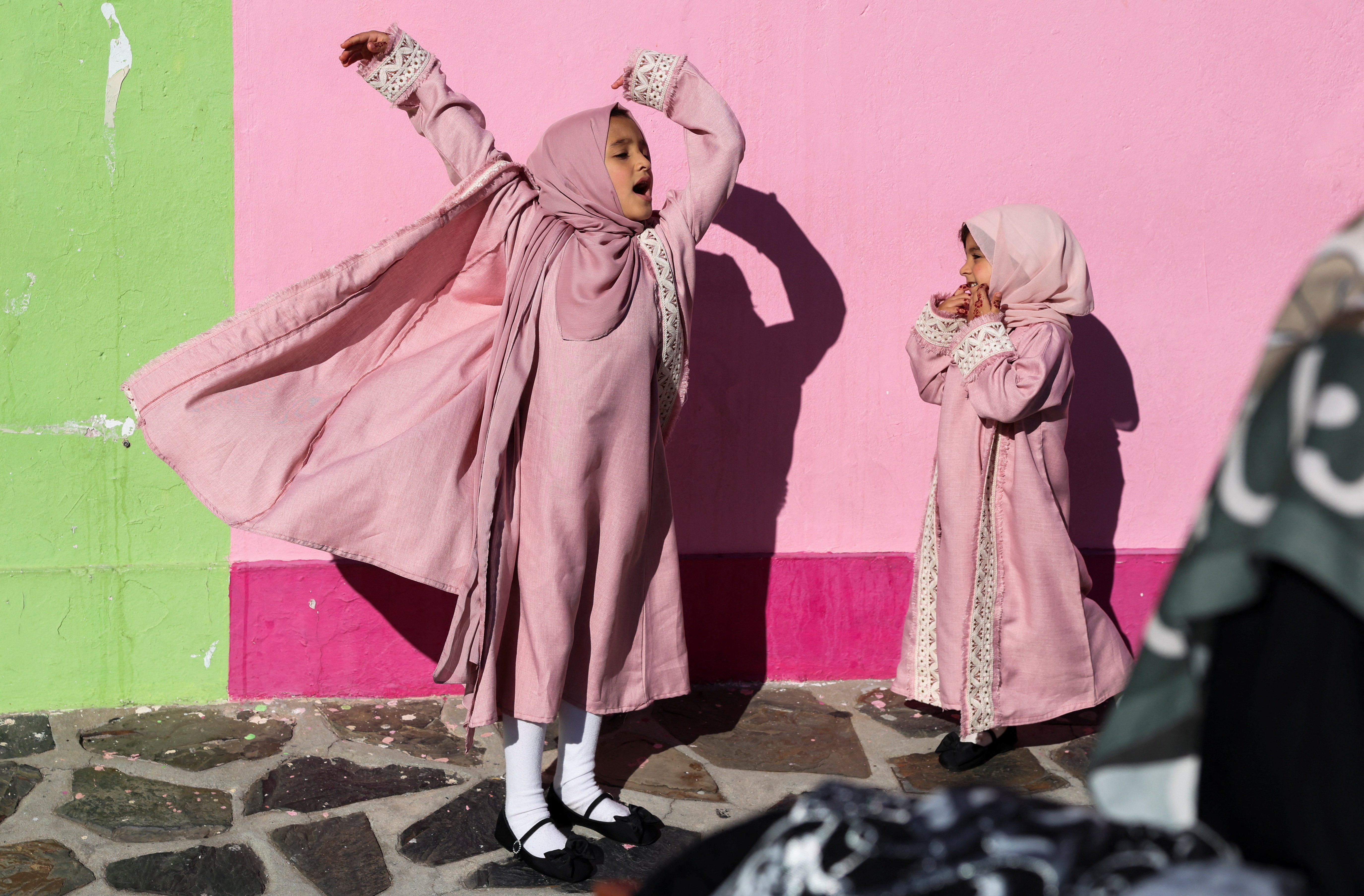 Two girls in colorful robes stand beside a pink painted wall.