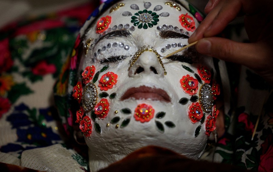 A bride has her face painted white and decorated with sequins while lying down.