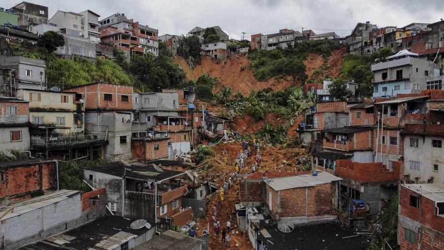 A wide view of a hillside neighborhood, partially wrecked by a mudslide, with rescue workers standing atop the debris and mud.