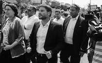 A group of individuals in a street, flanked by camerapeople