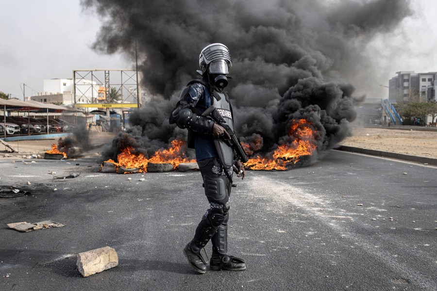 A riot police officer stands in a street beside a pile of burning tires.