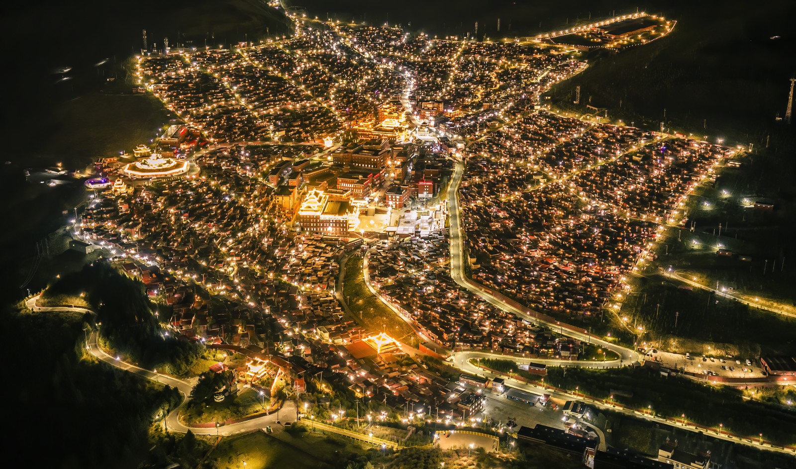 A nighttime aerial view of a collection of illuminated cabins situated around a larger Tibetan building