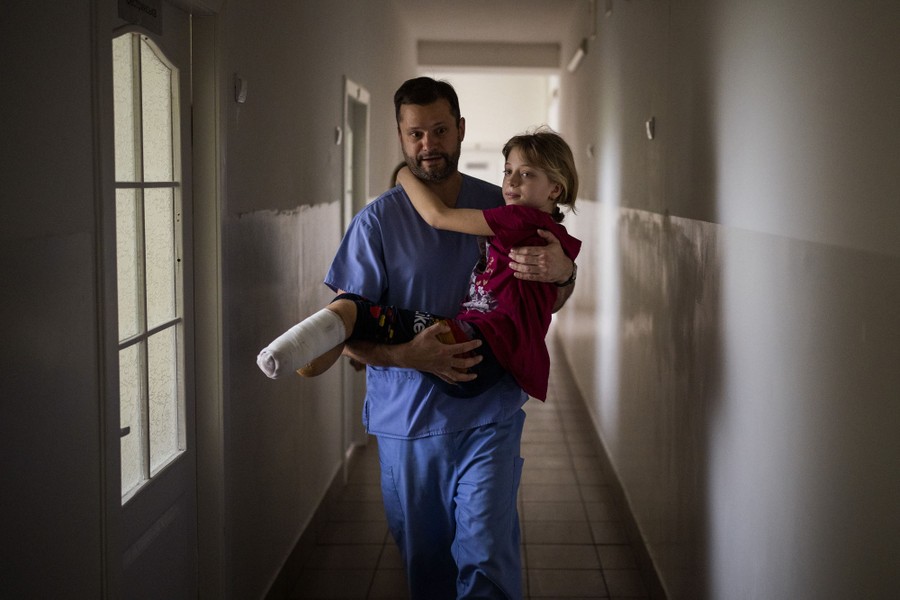 An injured child with a bandaged leg is carried by a doctor through a hospital hallway.