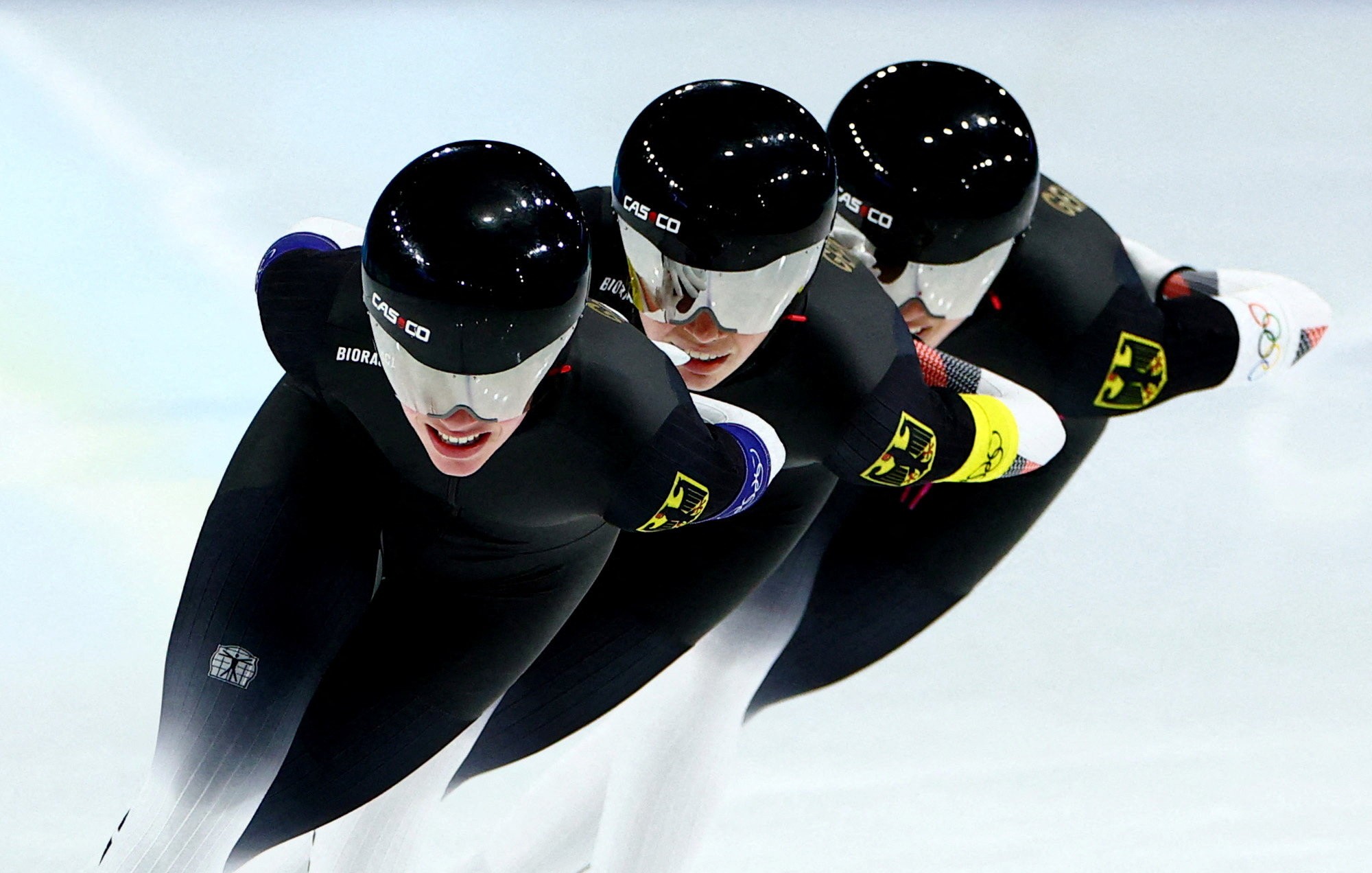 Three speed skaters race, seen close together wearing identical uniforms and helmets with mirrored visors.