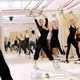 photo of reflection in mirror of Anderson leading a fitness class with both arms raised above head and hands holding weights in well-lit studio