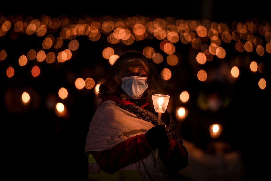 A pilgrim wearing a face mask holds a candle, as dozens of other candle flames are visible in the background.