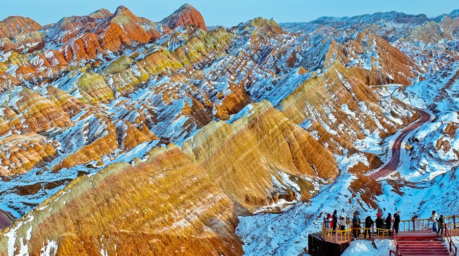 A small group of tourists stands in a viewing area overlooking a dramatic set of partially snow-covered striped mountains.