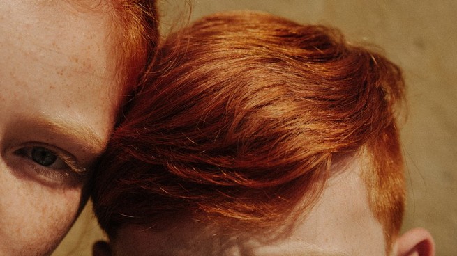 Ginger hair on the head of a young boy.  The corner of an older girl's face leans against him;  his eye is in the frame and looking at the camera.