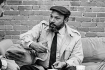 A black-and-white photo August Wilson in a trench coat, driving cap, and tie sitting on sofa and gesturing in conversation with a brick wall behind