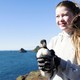 A young person holds a juvenile puffin while standing atop a sea cliff.