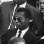 James Baldwin, seated among other people, wearing sunglasses and a suit and tie.