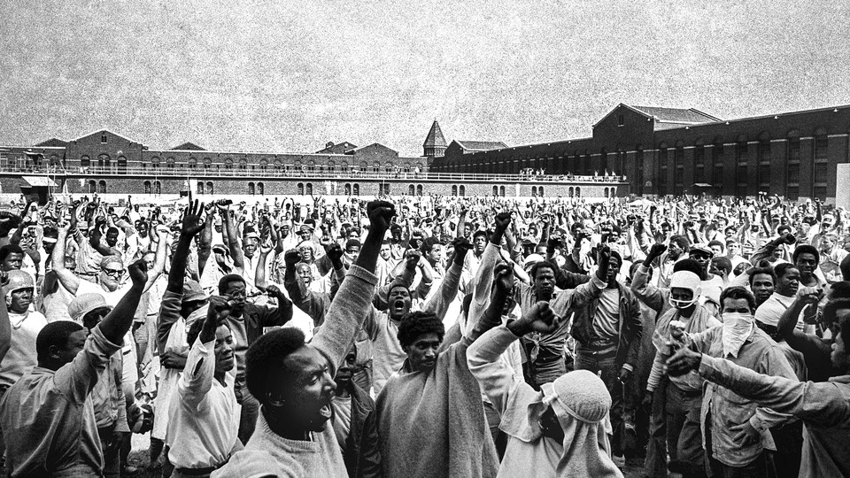 A black and white photograph depicts hundreds of inmates in the interior yard of Attica prison. Many have their hands raised in the "Black Power" gesture.