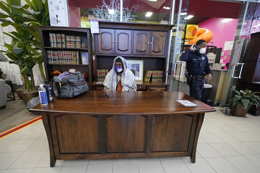 A man wearing a small blanket over his head sits at a floor display desk with displays of office furniture behind him, while sheltering in a furniture store.