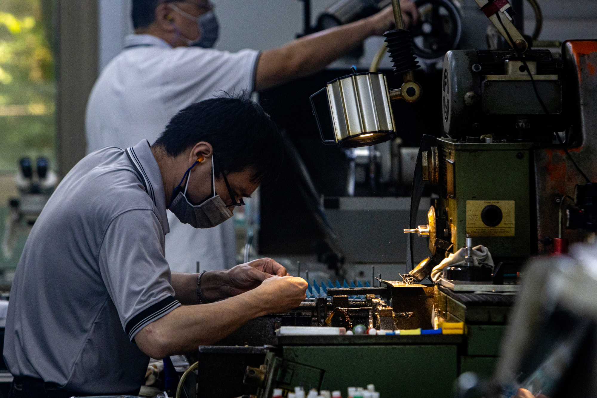 photo of man in glasses and mask sitting at cluttered workbench assembling electronics, with other workers in background
