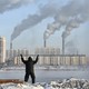 A photograph of a person facing away from the camera, standing with their arms raised, in front of factories with smokestacks emitting smoke
