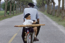 An old man and a young boy, both in berets, ride a bicycle with two baguettes strapped to the back down a tree-lined street; the young boy is looking back over his shoulder at the camera.