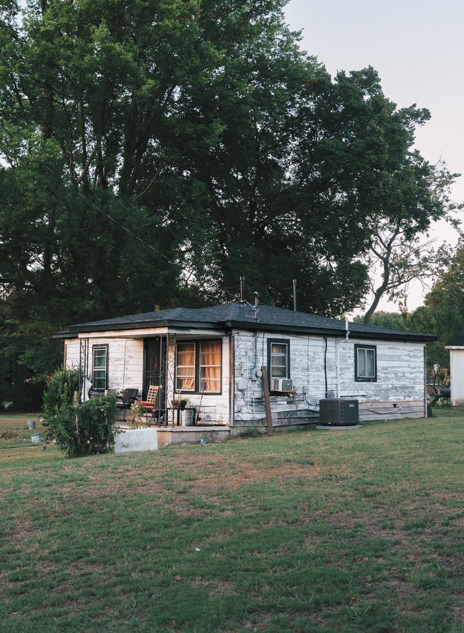 photo of simple railroad-style house with peeling white paint and large trees in background
