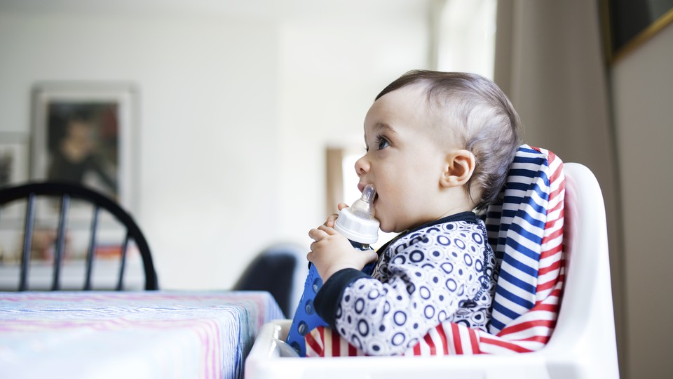 A baby sits in a high chair holding a bottle.