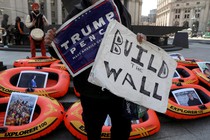 A person holding a "Trump, Pence" support sign and a poster reading "Build the Wall" stands in front of rafts meant to display opposition to the U.S. refugee ban in New York City.