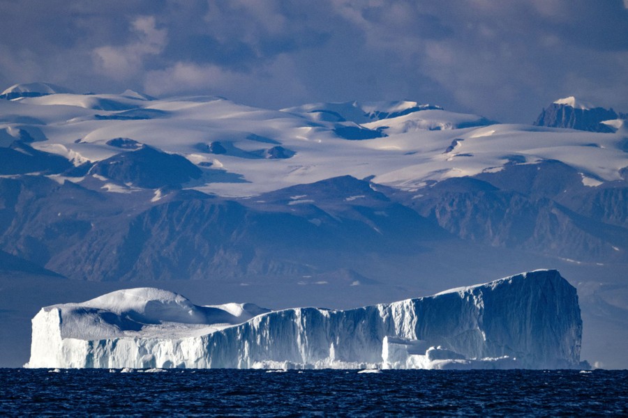 An enormous iceberg drifts along a mountainous coastline.