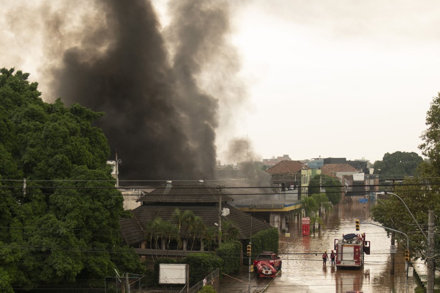 Black smoke rises above buildings, with a fire truck parked in a flooded street nearby.