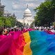 A large rainbow flag draped over a street full of parade marchers, with the U.S. Capitol Building in the background.