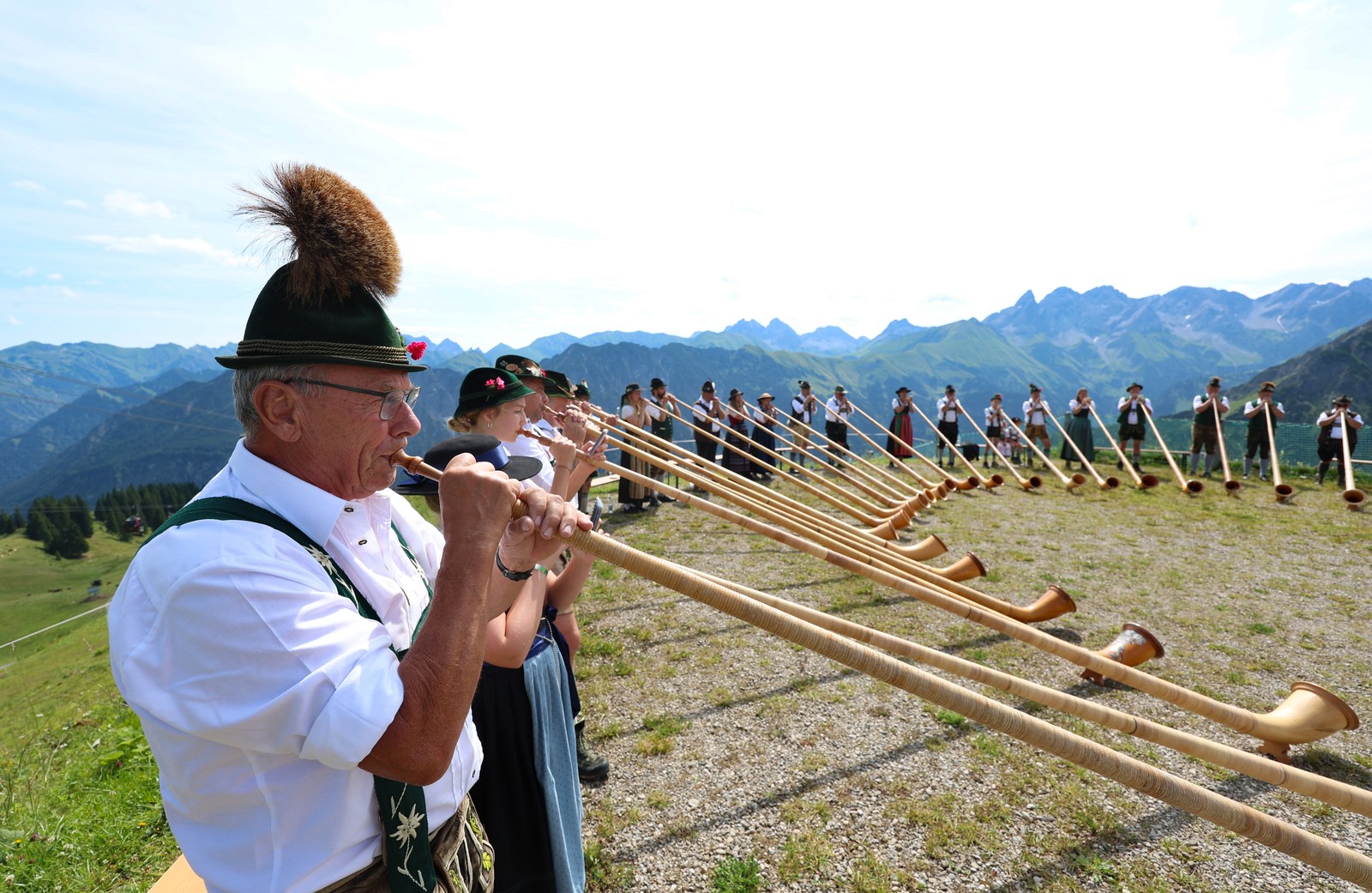 Dozens of people in traditional costume stand in a semicircle, playing alphorns.