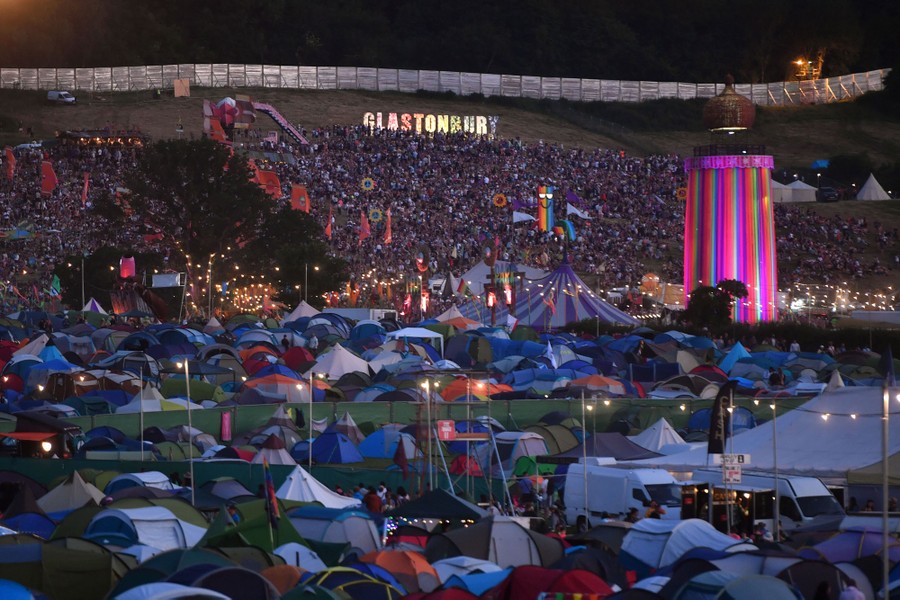 A field is covered with camping tents, in front of a crowd of people, with letters spelling out "Glastonbury" in the background.