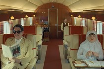 A color still from "The Phoenician Scheme" showing Benicio del Toro and Mia Threapleton in an airplane