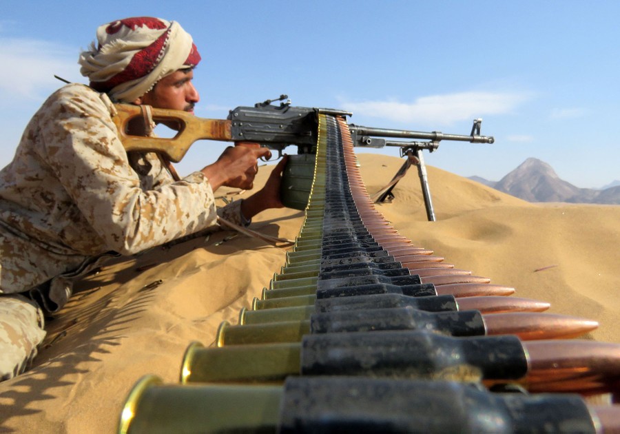 A fighter leans into his automatic weapon set on a bipod on a sand dune, a belt of ammunition running down toward the camera.