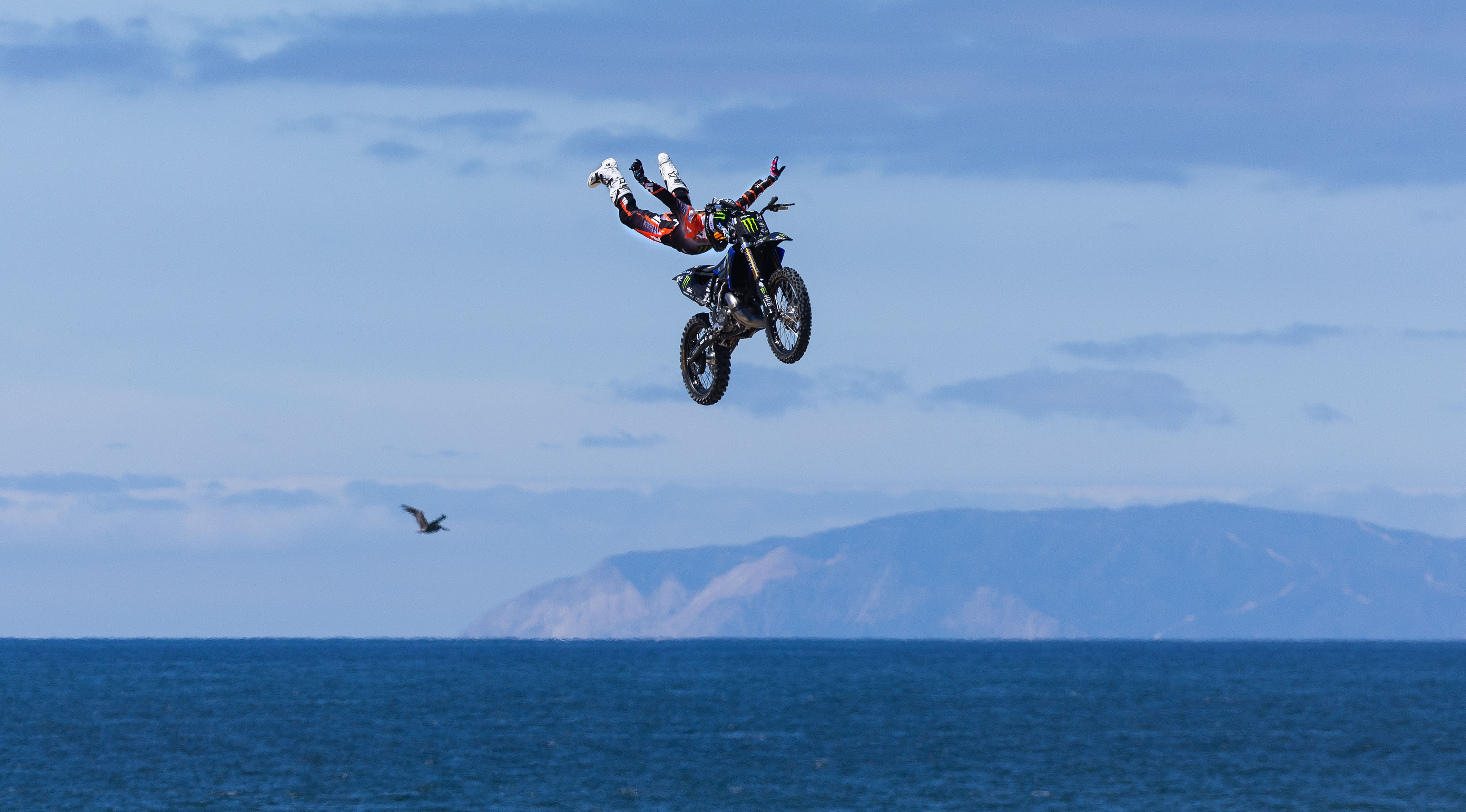 A motorcycle stunt rider extends their arms and legs during a jump, with the ocean visible in the background.