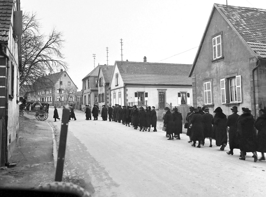 Dozens of mourners dressed in black walk through a a village street, following a horse-drawn hearse.
