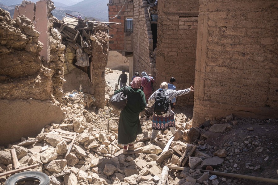 Six people walk carefully over piles of rubble, past damaged buildings.