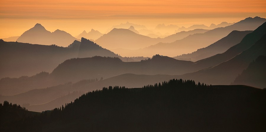 A view of a mountainous landscape, many peaks shrouded in a gentle fog, warmly lit by a low sun.