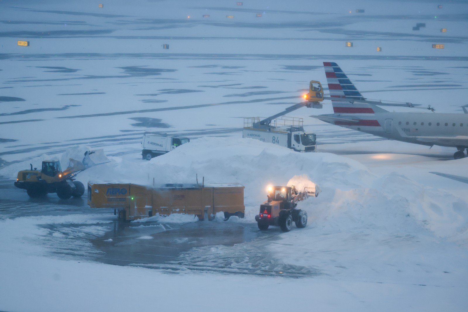 Two front-end loaders dump snow into an ice melter near a passenger aircraft.