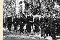A black and white photograph of male Harvard students from the 1950s in graduation robes processing past an ivy-lined building and the John Harvard statue