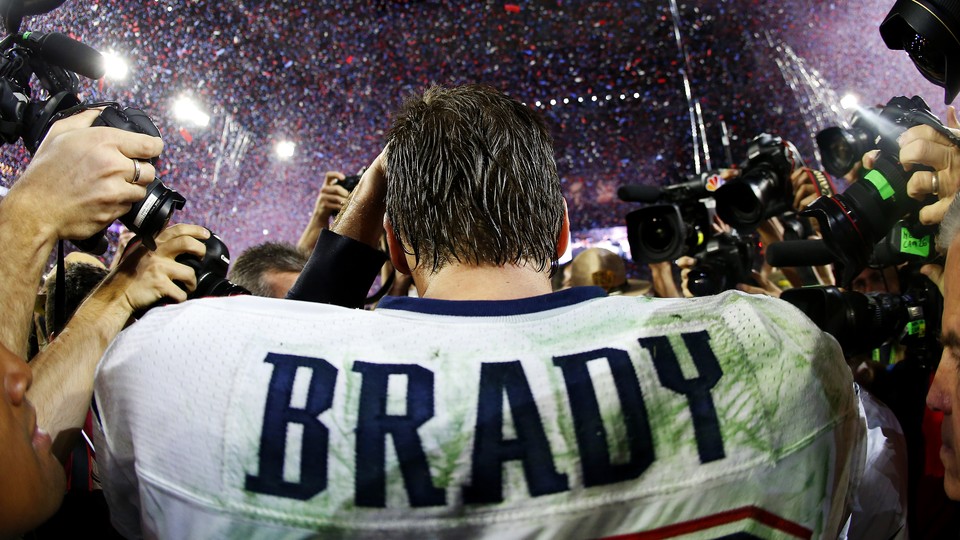 Tom Brady walks away from the camera in a football stadium, as a horde of photographers takes pictures of him.