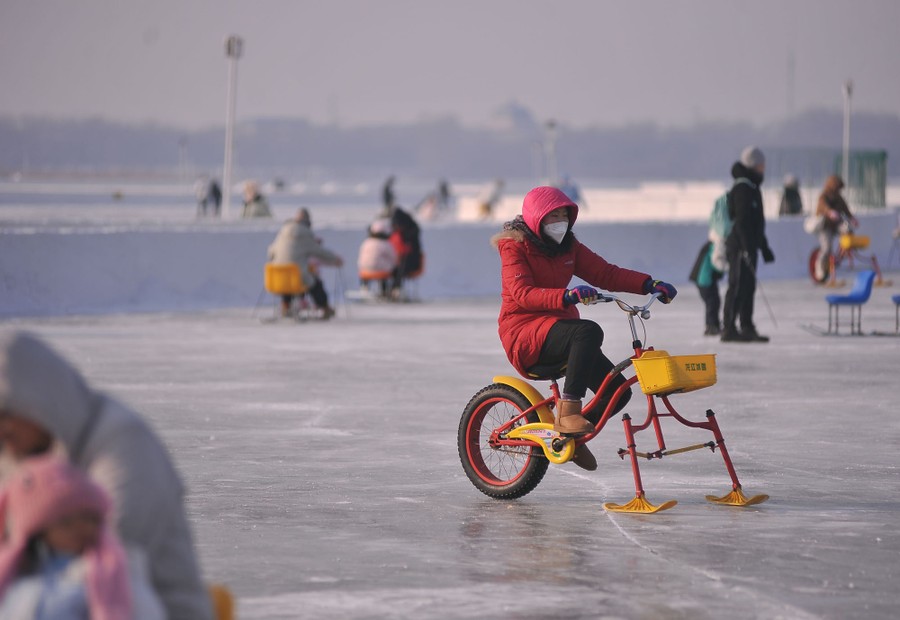 People ride on a frozen lake, using chairs with skis, and a modified bicycle with two skis in place of its front wheel.
