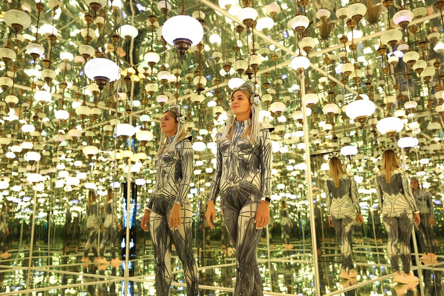 A person wearing a futuristic costume walks through a room of mirrors and lights.