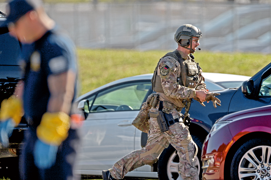 photo of man in full camouflage military-style gear with helmet, ballistic vest, and holstered weapon running past parked cars while pulling on gloves