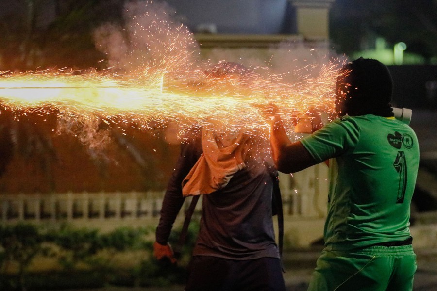 Two protesters stand in a street, as one launches some sort of firework from a tube.
