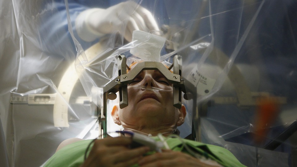 A patient lies on an operating table in front of a plastic sheet, behind which a person in scrubs and gloves performs surgery on the patient's brain.