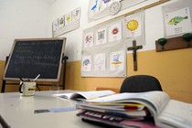 A teacher's desk is covered with papers in a classroom with a chalkboard and crucifix.