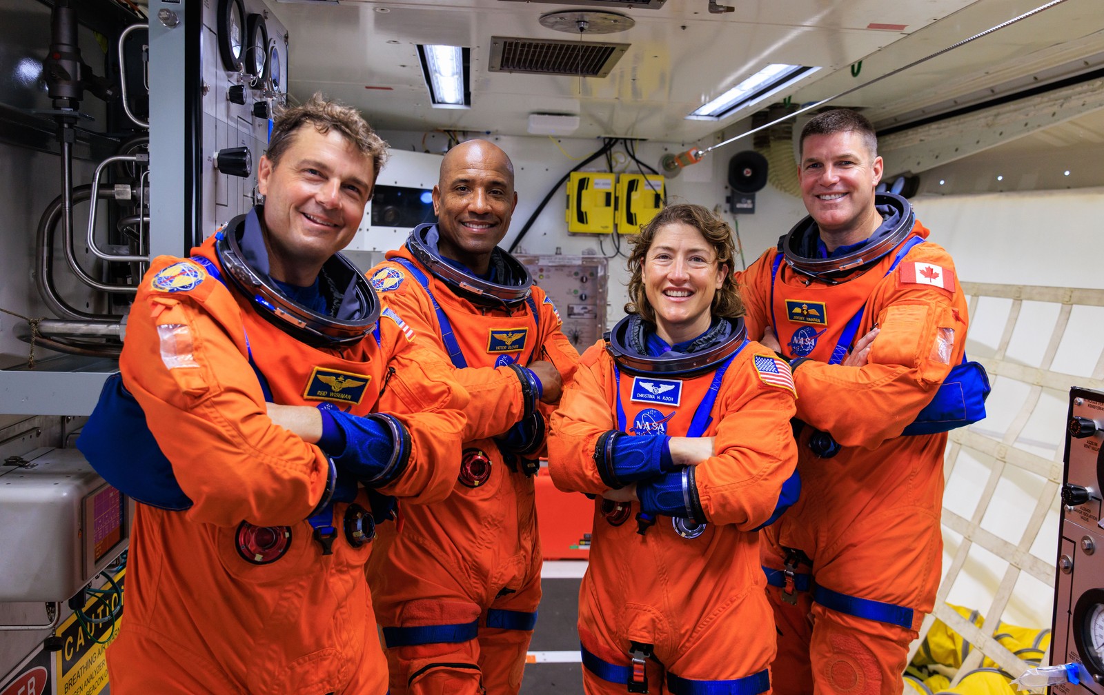 A crew of four astronauts stand, posing in flight suits, inside a small white room.